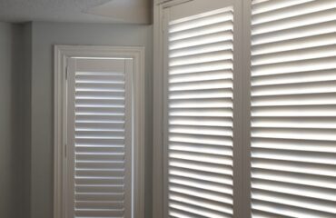 why-wood-shutters-remain-a-timeless-choice-for-every-home Two white wood shutters frame the windows in a room, letting in filtered sunlight.